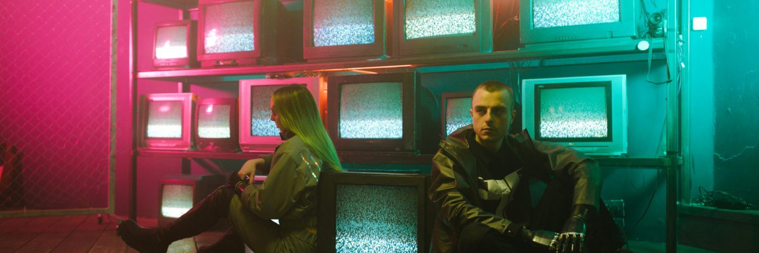Two individuals in a cyberpunk setting with neon lighting and stacked television screens.